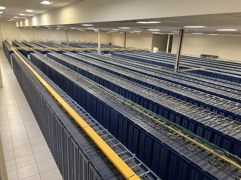 Long rows of blue cabinets under cable trays in a datacenter hall.